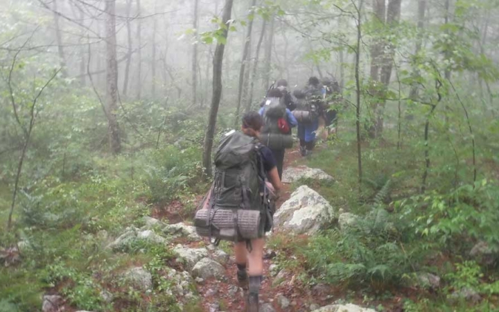 a group of young people wearing backpacks hike away from the camera through a foggy wooded area 